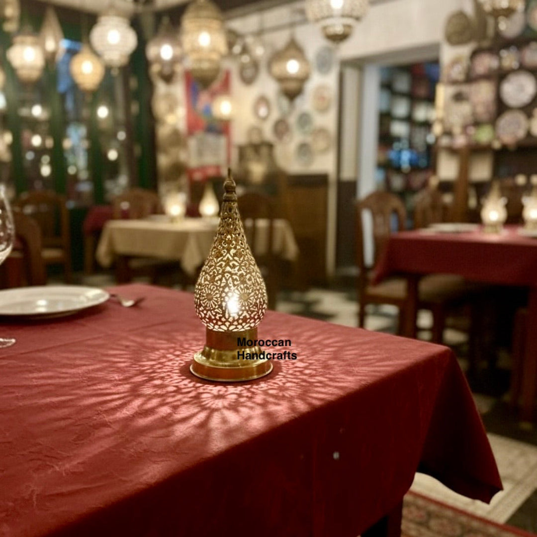 Traditional Moroccan dining ambiance featuring a handcrafted brass lantern on a rustic wooden table. The metalwork casts detailed arabesque shadow patterns, complementing the red patterned cushions and the dark, richly tiled walls of the restaurant.