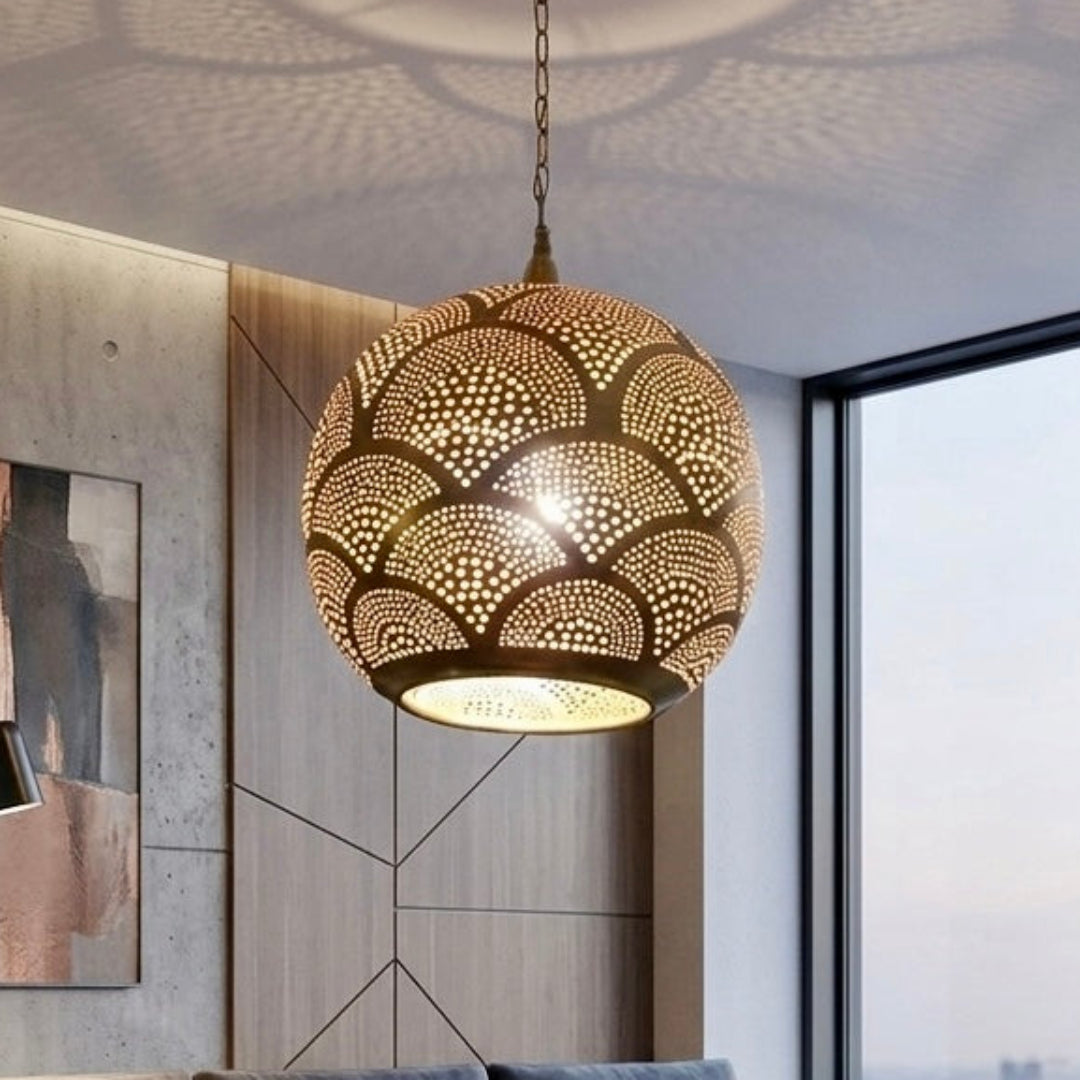 Large handcrafted Moroccan brass globe pendant lamp hanging in a modern living room with concrete walls and a grey sectional sofa, casting scalloped light patterns.