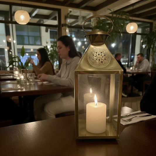 A close-up, low-light shot of a lit white pillar candle inside a decorative brass lantern centerpiece on a dark wooden restaurant table. In the softly lit background, patrons are seated and dining, suggesting a cozy, intimate atmosphere.