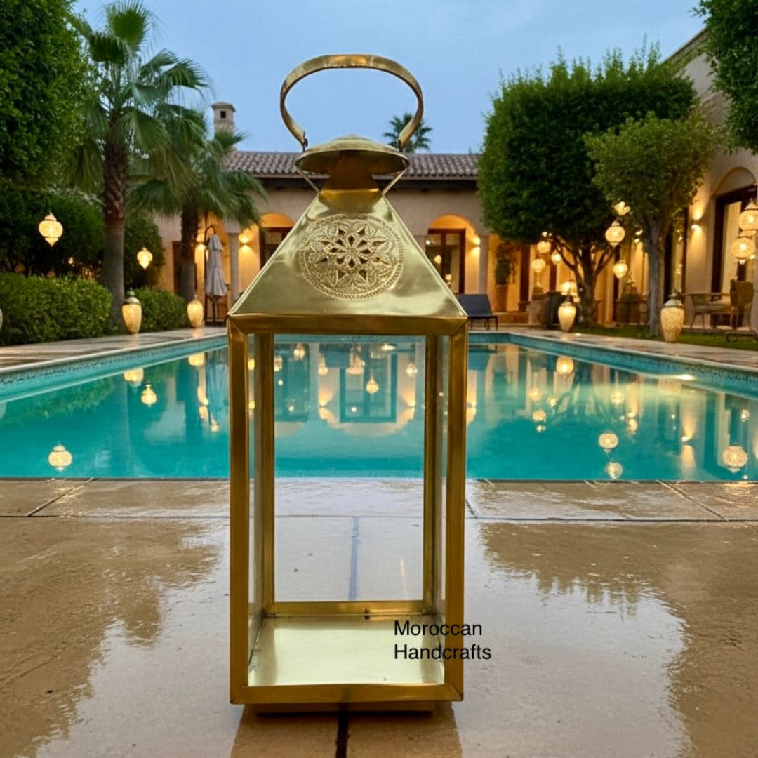 Vertical view of a traditional square Moroccan-style lantern with clear glass sides, made of polished brass, providing warm lighting in a dining environment.