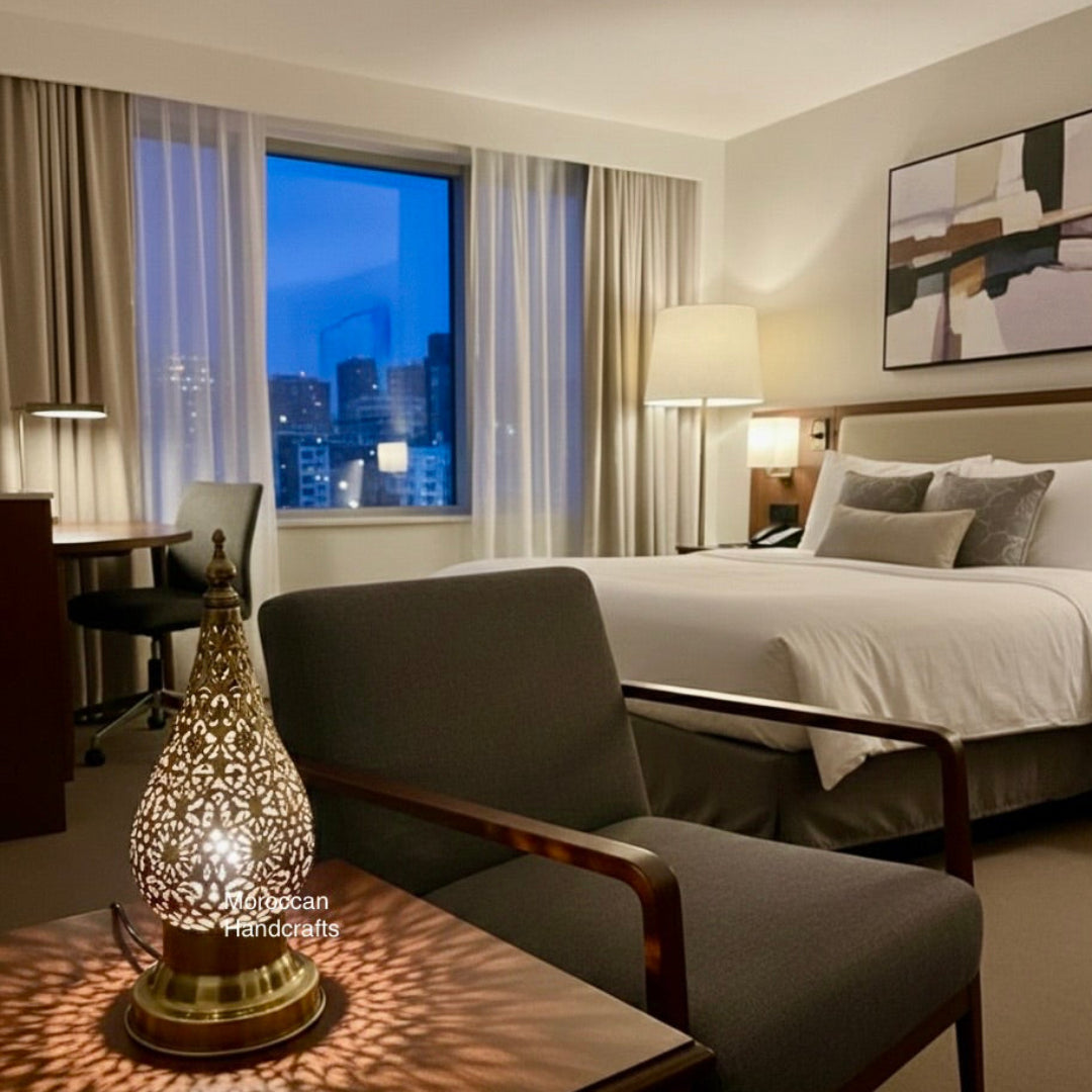 Handcrafted brass Moroccan lantern (or table lamp) with a cone shape and detailed metalwork, illuminating a table setting with a small tagine and two glasses of mint tea. The lamp's warm glow and shadows create a cozy, intimate atmosphere.