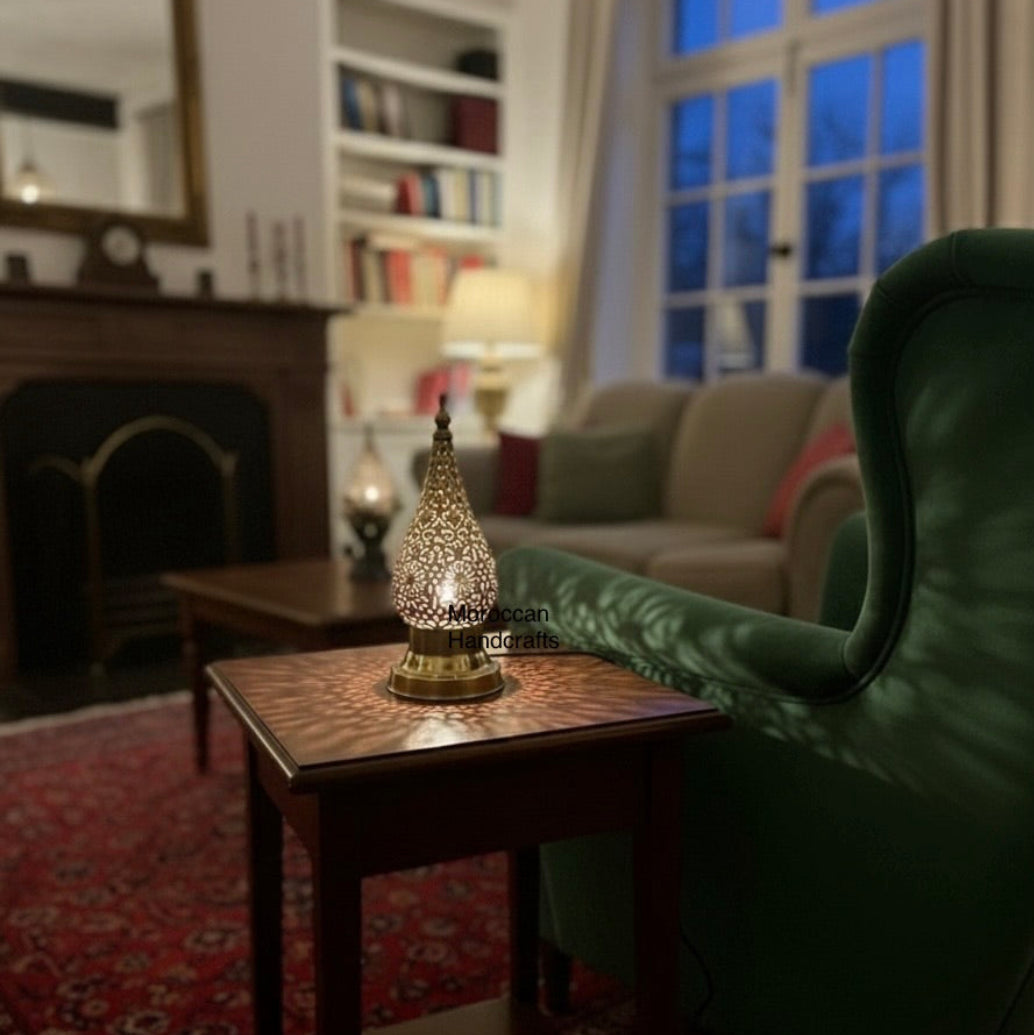  Traditional Moroccan dining ambiance featuring a handcrafted brass lantern on a rustic wooden table. The metalwork casts detailed arabesque shadow patterns, complementing the red patterned cushions and the dark, richly tiled walls of the restaurant.