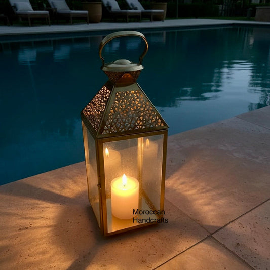 A tall, gold-finish Moroccan brass lantern with an intricate filigree top, featuring a lit white pillar candle on a wooden restaurant table.