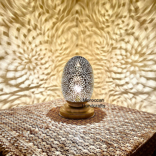 Moroccan-style brass table lamp in an egg shape with intricate filigree, casting a magnificent shadow pattern on the wall and surface next to the lamp.