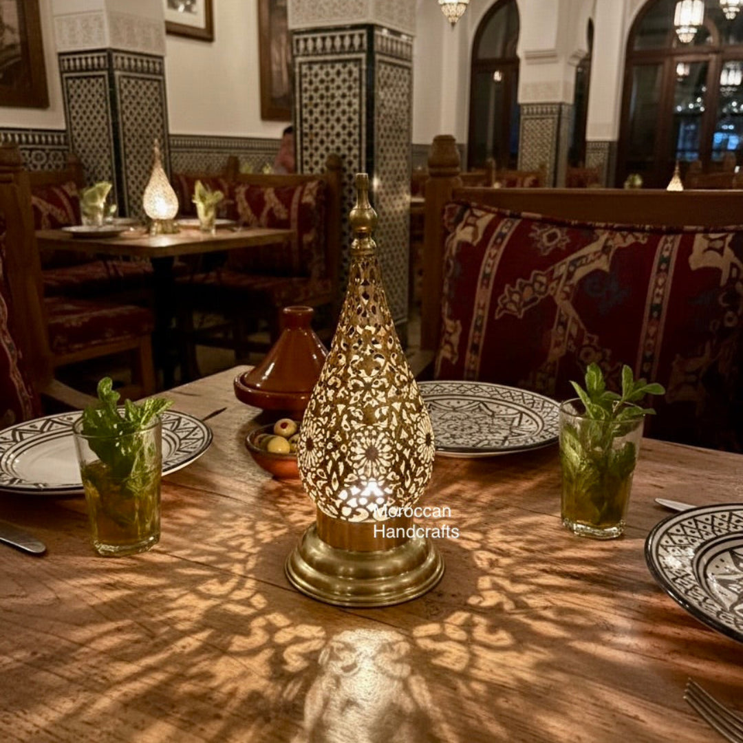  A close-up shot of a dimly lit Moroccan restaurant table. A tall, golden brass table lamp with intricate filigree patterns sits in the center, casting beautiful, ornate shadows onto the wooden tabletop. On either side are glasses with fresh mint leaves and a small Moroccan tagine dish.