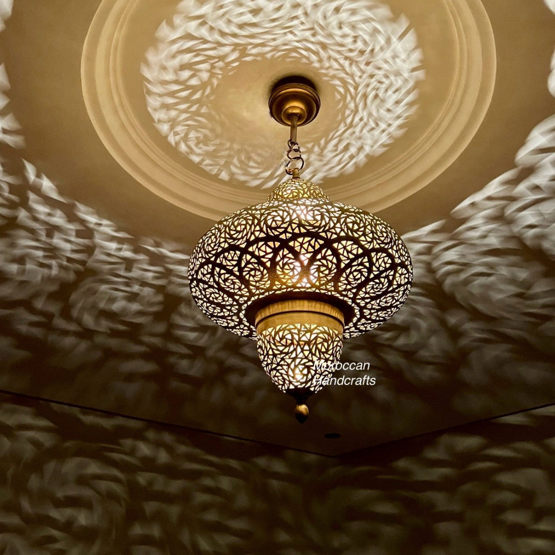  A close-up of a decorative Moroccan brass urn-shaped pendant lamp with detailed floral and geometric perforations. The light casts mesmerizing, swirling shadow patterns across the brown wall and ceiling medallion.