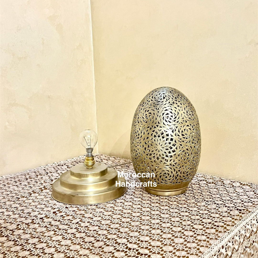 Detailed shot of a Moroccan brass egg-shaped table lamp with intricate, hand-pierced filigree, casting a beautiful, complex shadow pattern onto the wall and surrounding surface.