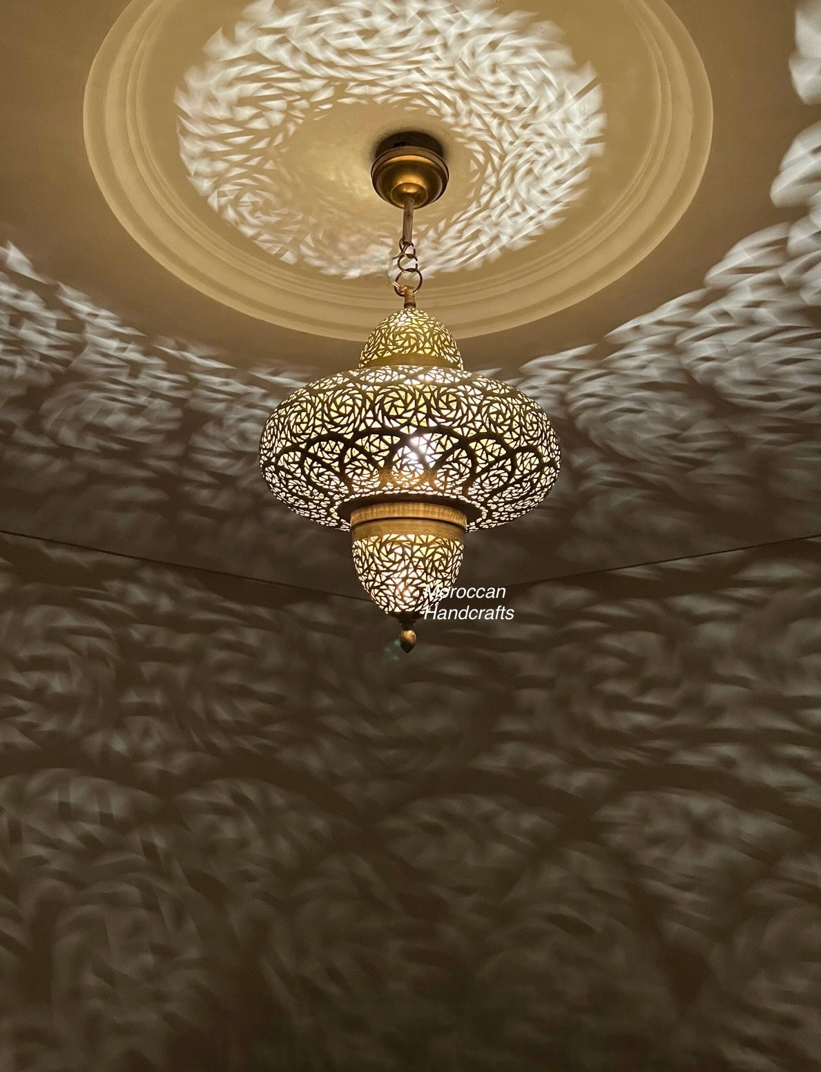 A large, spherical Moroccan brass pendant lamp with intricate, geometric filigree patterns. It hangs over a beige sectional sofa in a modern, neutral-toned living room, casting striking shadow patterns on the walls.