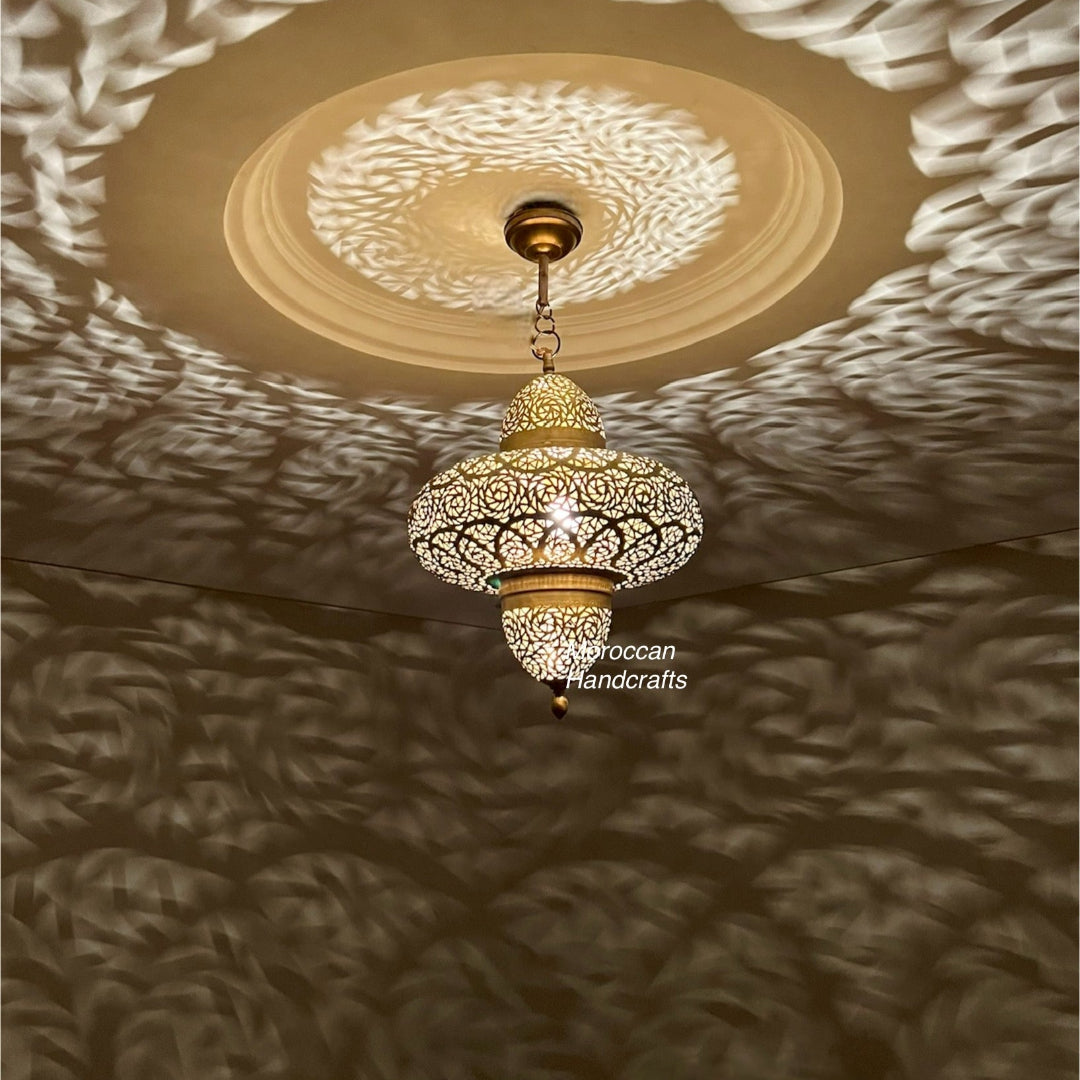 A large, spherical Moroccan brass pendant lamp with intricate, geometric filigree patterns. It hangs over a beige sectional sofa in a modern, neutral-toned living room, casting striking shadow patterns on the walls.