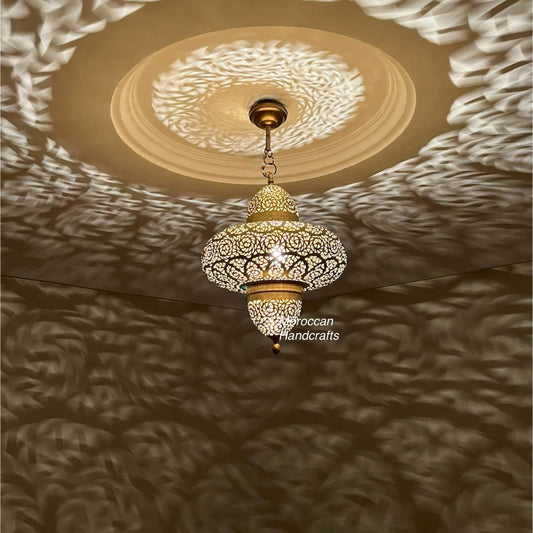 A large, spherical Moroccan brass pendant lamp with intricate, geometric filigree patterns. It hangs over a beige sectional sofa in a modern, neutral-toned living room, casting striking shadow patterns on the walls.