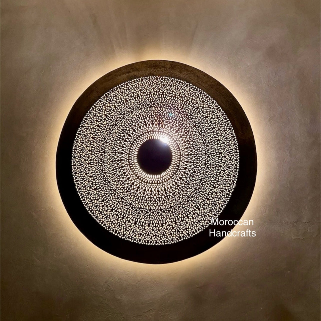 Moroccan handcrafted brass wall sconce light with intricate laser-cut or filigree geometric pattern. The round lamp casts a warm, decorative glow and stunning shadow patterns.