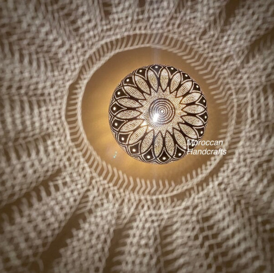 Compact oval/egg-shaped Moroccan table lamp made of hand-carved brass, casting a gentle, ambient swirling light pattern on a woven surface.