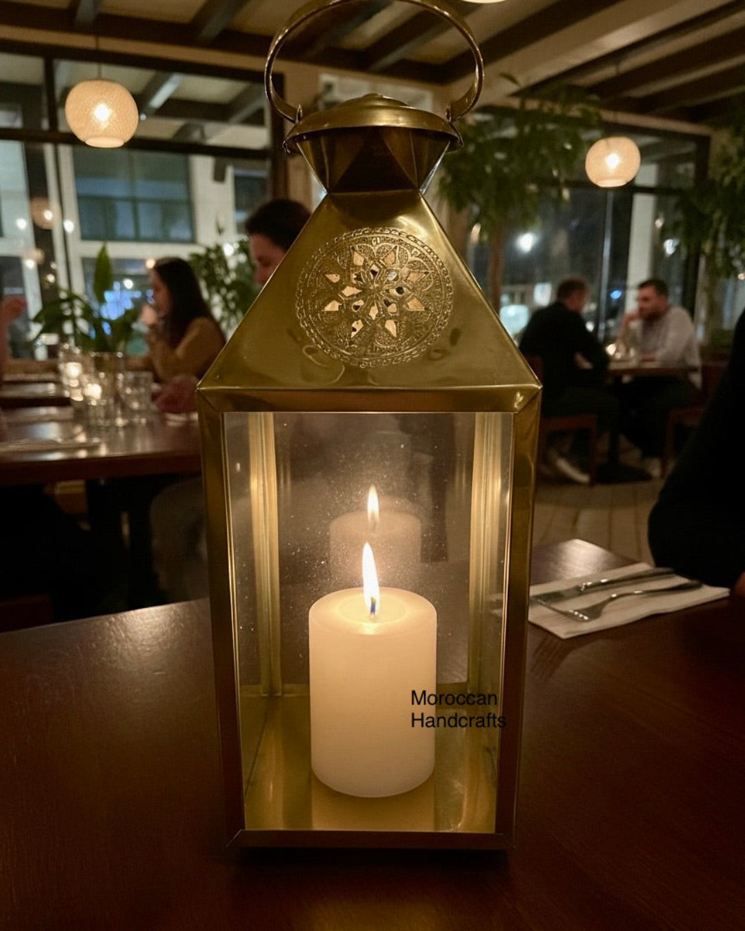 Handcrafted tall brass Moroccan lantern featuring ornate geometric perforations on the roof and a lit white pillar candle, set on a dark wood table.