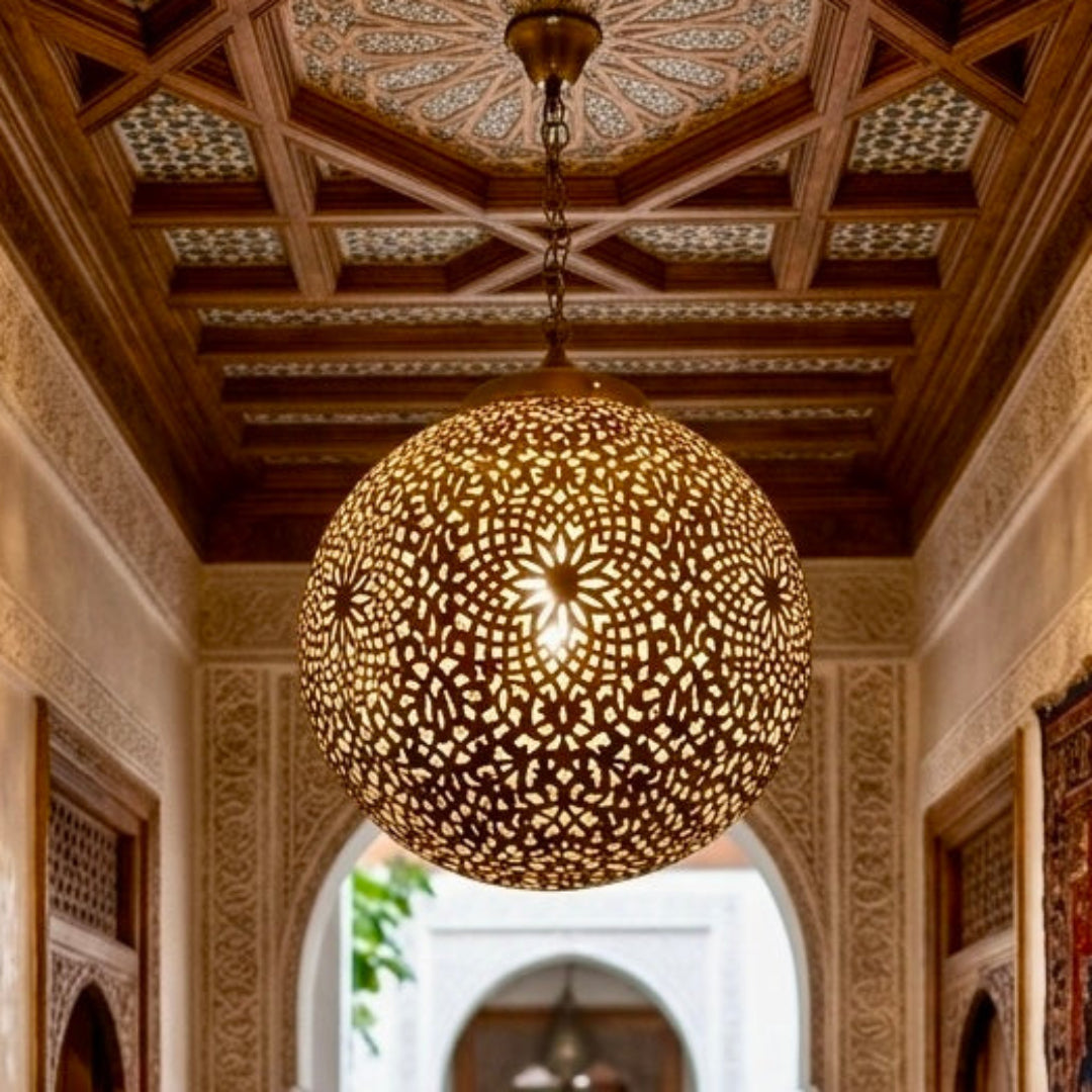 Large spherical Moroccan brass pendant light hanging in a traditional riad hallway with carved wood ceilings and red rugs.