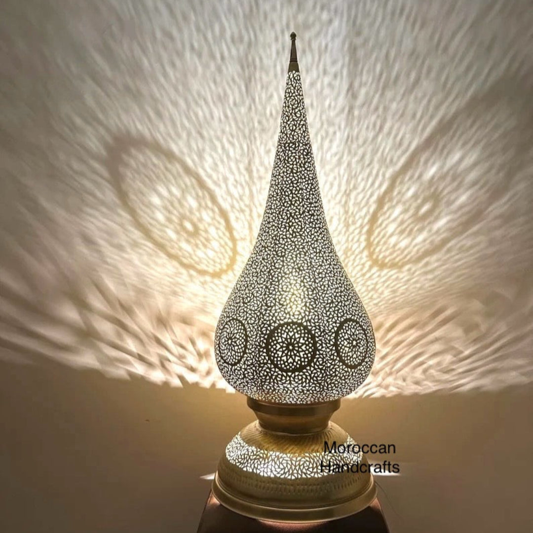 Tall Moroccan brass floor lamp in a tear drop or minaret shape, featuring detailed filigree and geometric patterns that cast large shadow hearts.