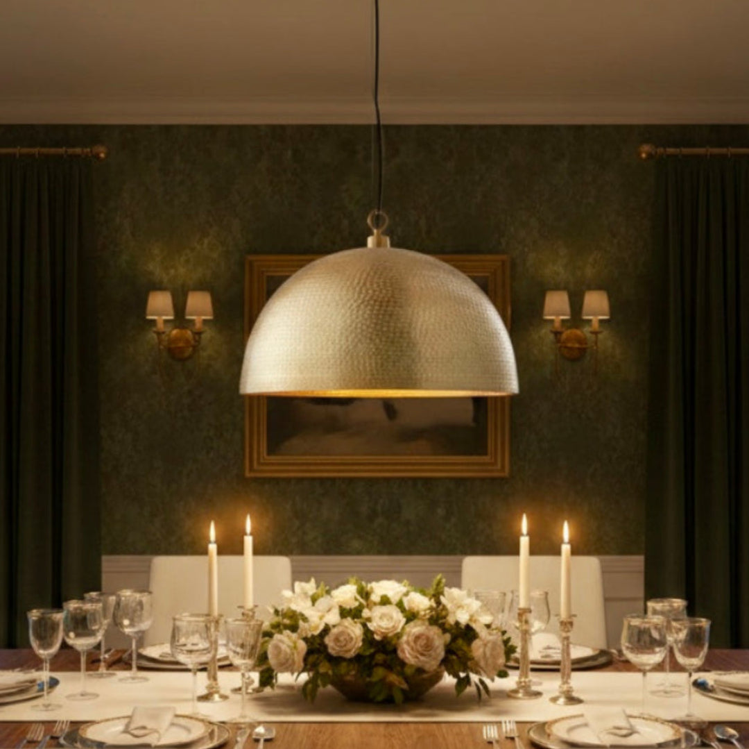 A large hand-hammered brass dome pendant light hanging over a formal wooden dining table with white candles and green floral wallpaper background.