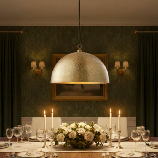 A large hand-hammered brass dome pendant light hanging over a formal wooden dining table with white candles and green floral wallpaper background.