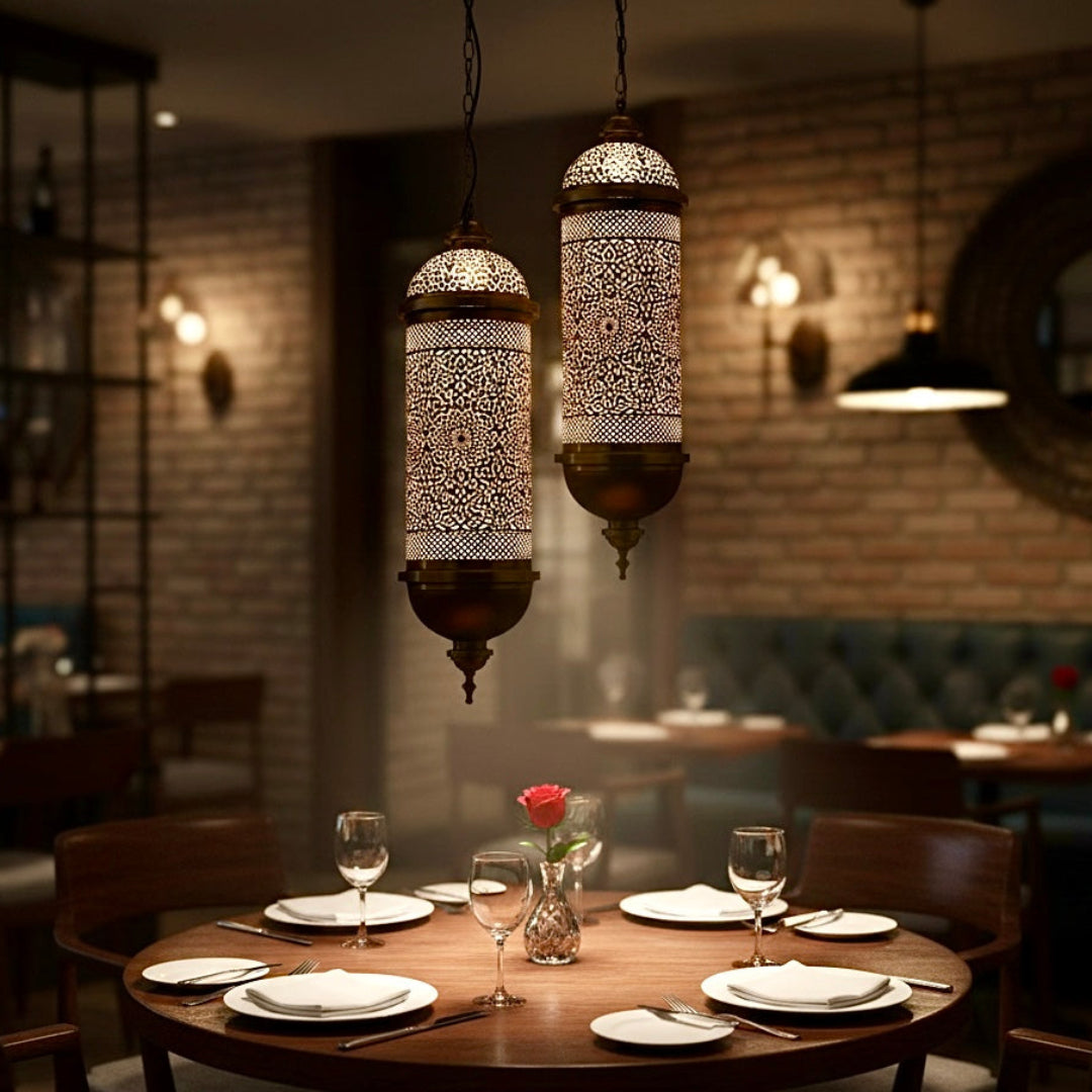  Two handcrafted brass Moroccan pendant lights hanging over a wooden dining table in a cozy, rustic restaurant setting.