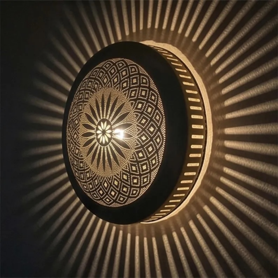 A close-up side angle of a black Moroccan flush mount brass lamp turned on, projecting dramatic radial geometric shadow lines across a warm wall.