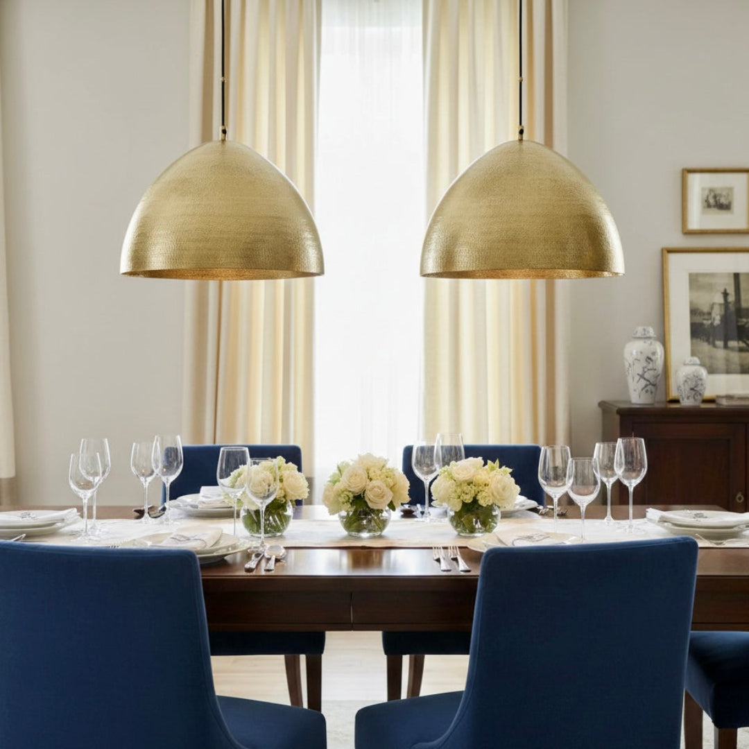 Two golden hammered brass dome lamps suspended above a formal dining table with blue chairs and white roses.