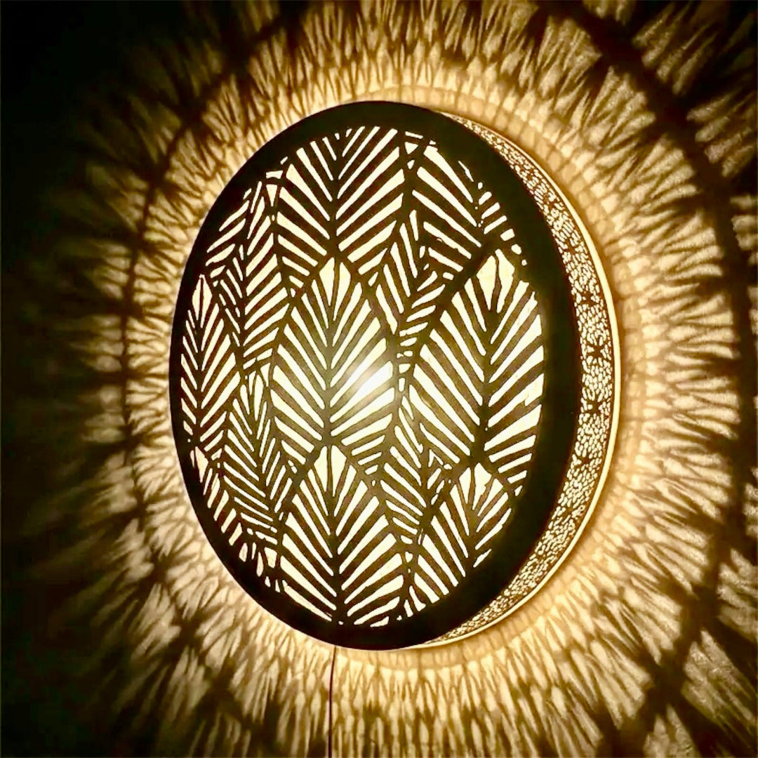 Close-up of a round Moroccan brass wall sconce illuminated, displaying a warm glow and precise geometric leaf-cutout shadow effects.