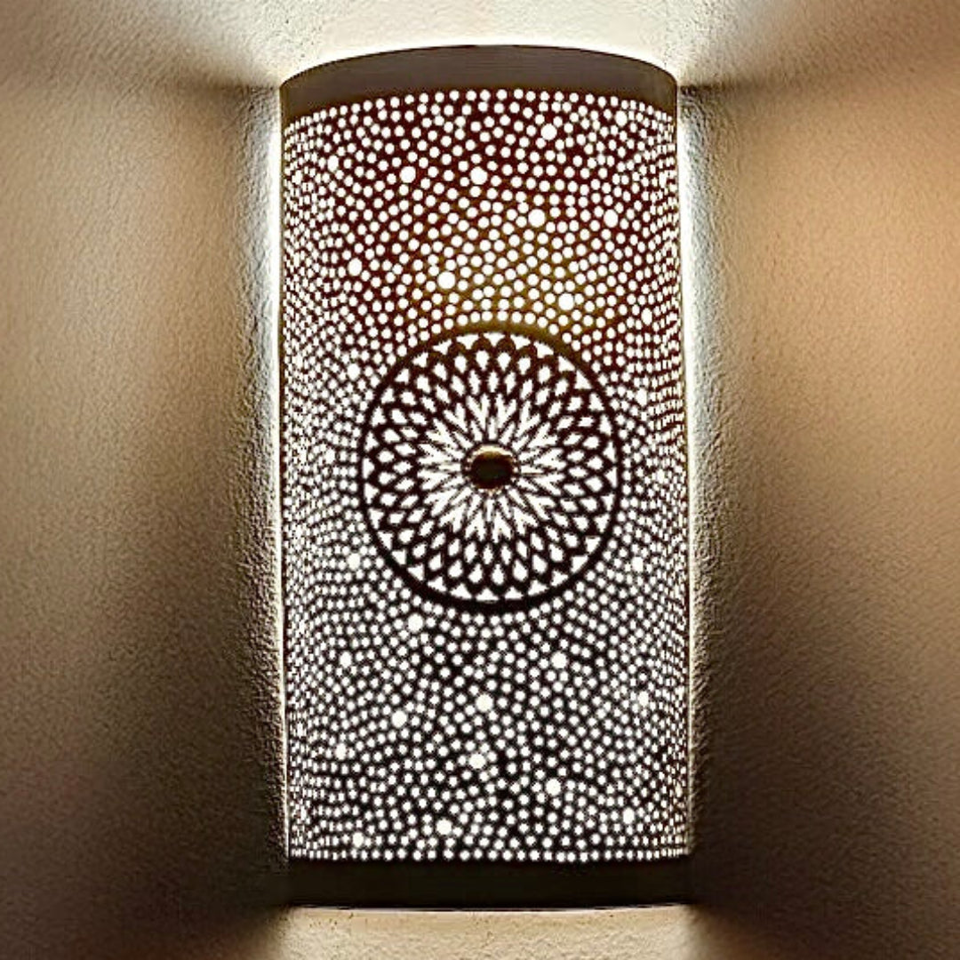  Close-up of a Moroccan handmade brass wall lamp illuminated, showing the detailed geometric flower pattern and light projection on the wall.