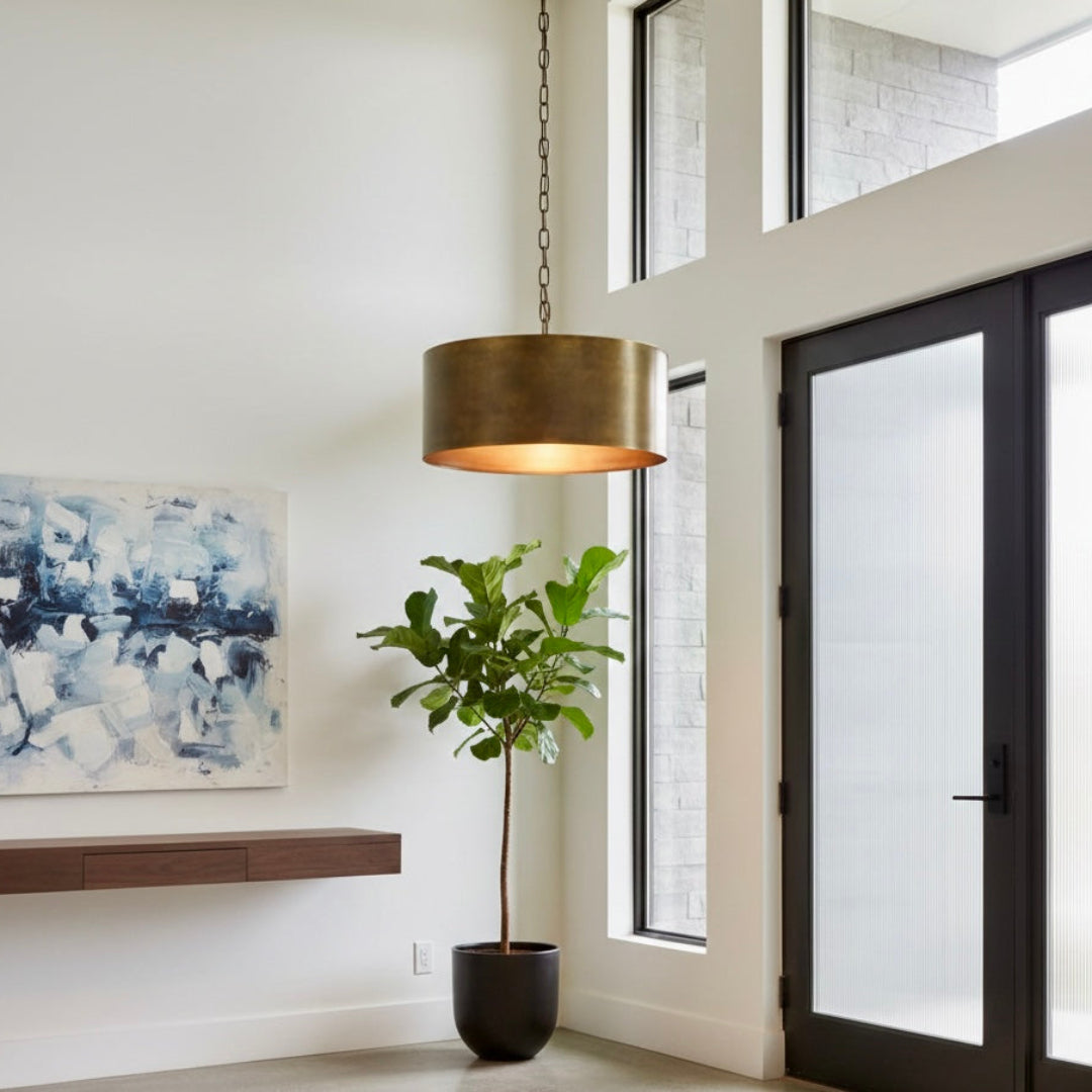 Modern brushed gold drum pendant light in a bright minimalist room with large windows and a fiddle leaf fig plant.