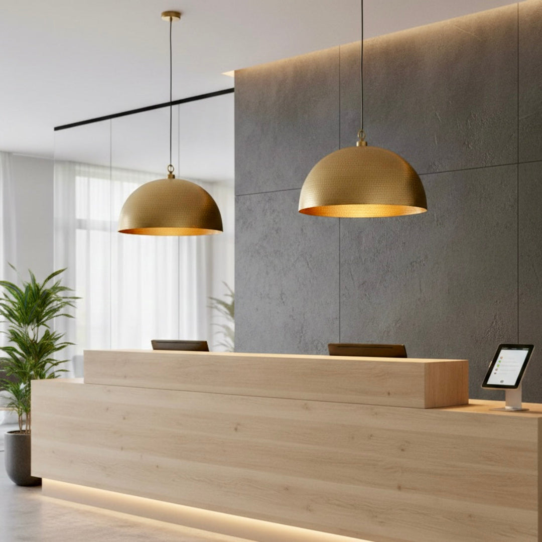 Pair of modern brass dome lamps hanging over a light wood reception desk in a bright, minimalist office setting with a grey concrete wall.
