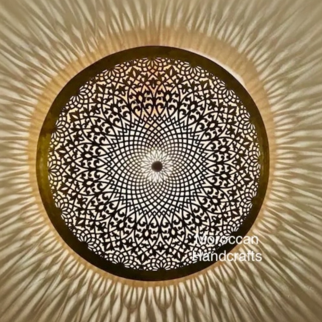 Fully illuminated Moroccan handmade brass ceiling fixture displaying its delicate openwork patterns and throwing dramatic radial shadow rays across a contemporary room ceiling.