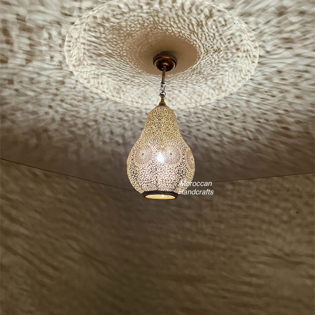 Handcrafted Moroccan brass pendant light hanging above a bed in a modern neutral bedroom, casting intricate shadows on the ceiling.