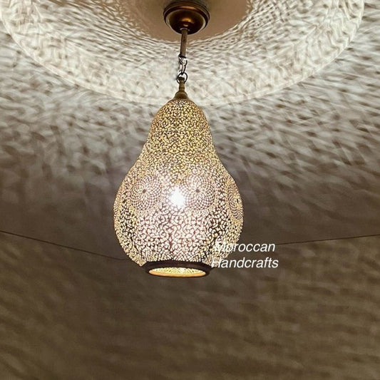 Handcrafted Moroccan brass pendant light hanging above a bed in a modern neutral bedroom, casting intricate shadows on the ceiling.