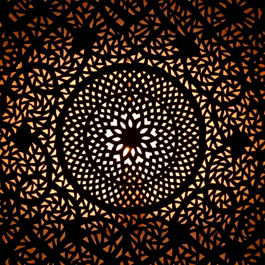 Extreme close-up of the detailed geometric hand-pierced metalwork pattern on a Moroccan brass lamp with light shining through.