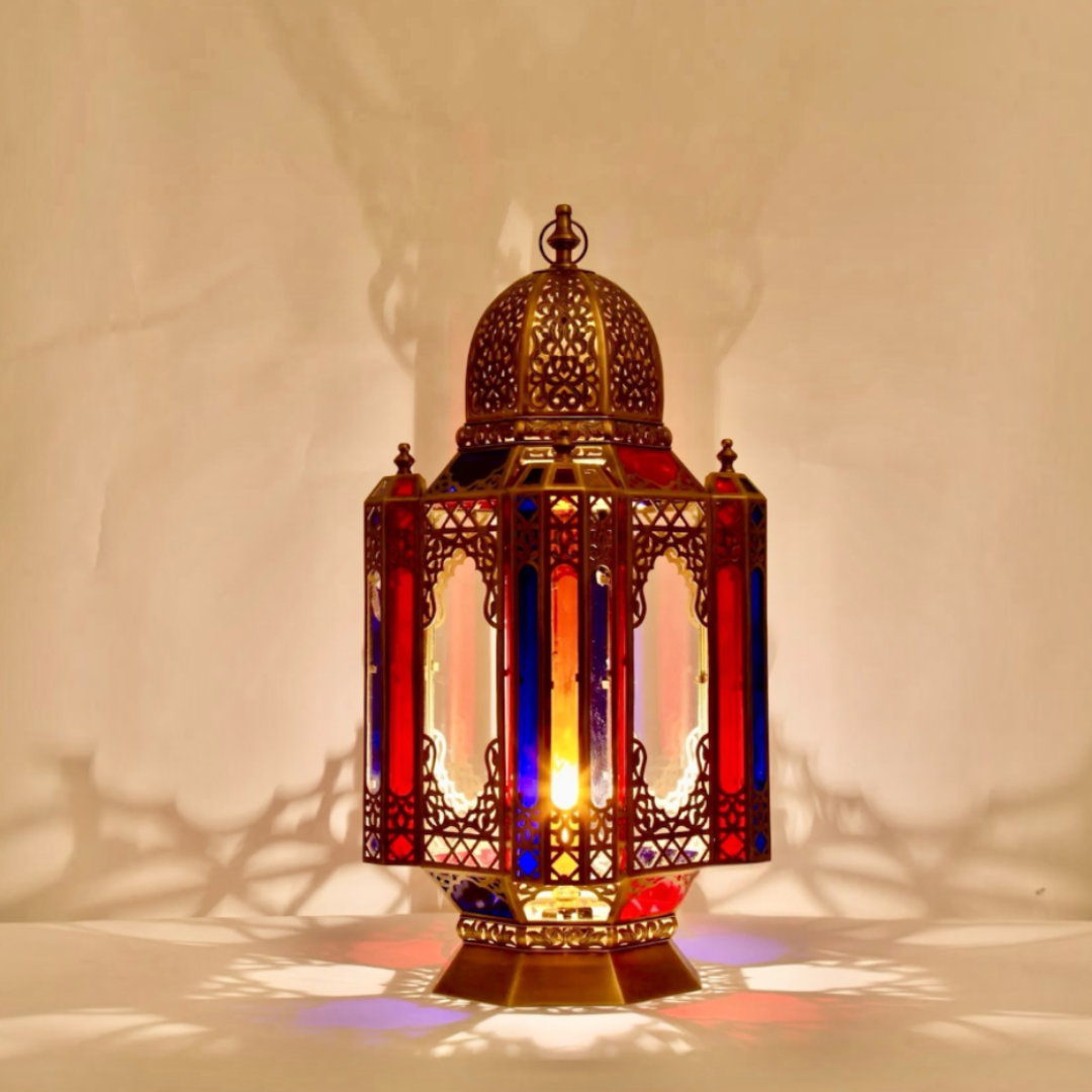 Lit Moroccan stained glass table lantern handmade from brass casting warm colorful shadows on a neutral wall.