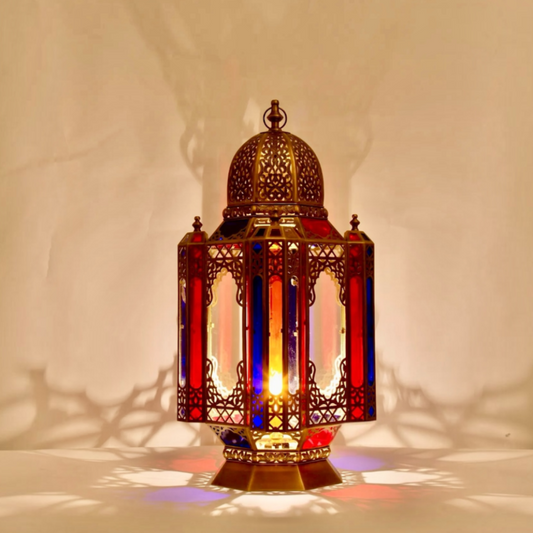 Lit Moroccan stained glass table lantern handmade from brass casting warm colorful shadows on a neutral wall.